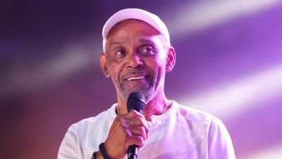 Maze's lead vocalist, Frankie Beverly, interacts with the crowd while wearing a smart suit and performs live on stage with a microphone.